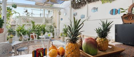 A tropical outdoor dining area with a wooden table, chairs, and a variety of fruits including pineapples and mangoes.
