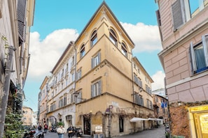 A narrow street with a corner building featuring a balcony and a shop with outdoor seating. - PIAZZA NAVONA WELLNESS LUXURY APARTMENT 8 PEOPLE (Roma)