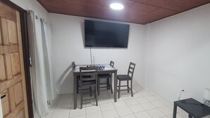 A room with a wooden ceiling, a flat-screen TV mounted on the wall, a dining table with chairs, a small table with a lamp, and a door with curtains. - Wonderful 1-bedroom apartment with AC in welcoming Noord (Noord)