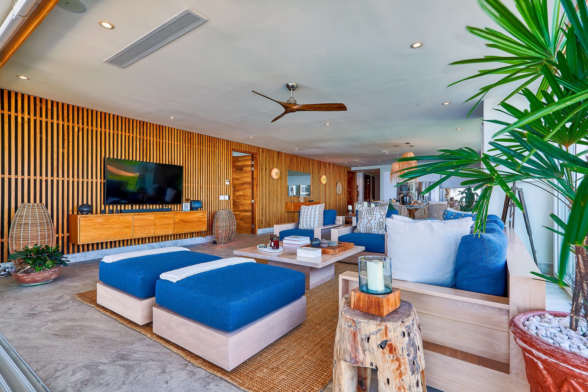 A modern living room with a large TV, blue sofas, wooden paneling, and a ceiling fan.