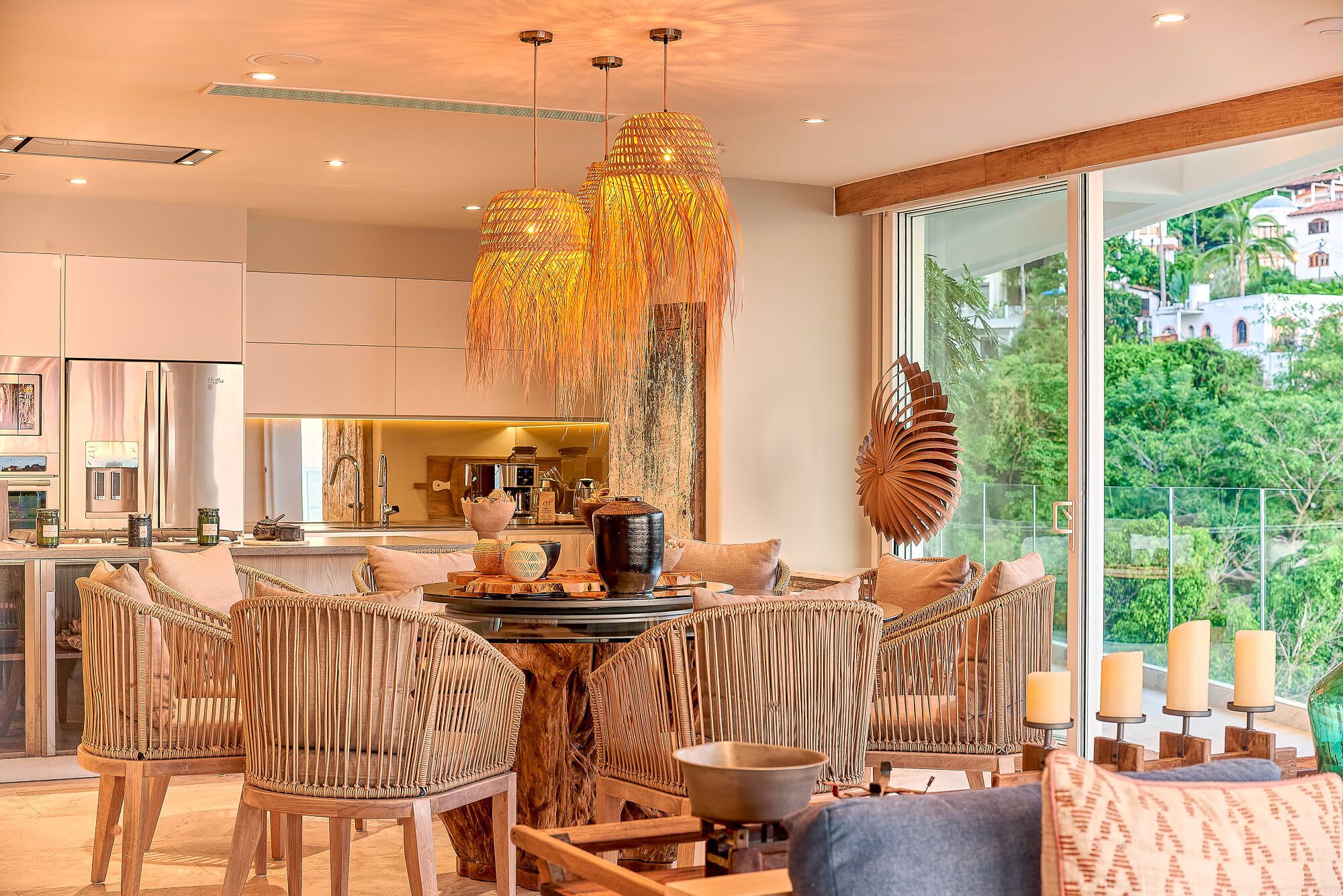 A modern kitchen with stainless steel appliances, a dining area with wicker chairs, and a large window offering a view of greenery.