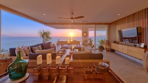 Amplia sala de estar con televisor grande, muebles de madera y vista al mar al atardecer.