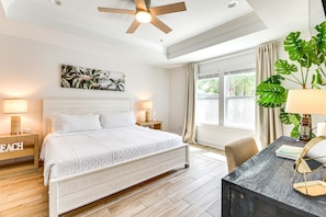 A bedroom with a bed, bedside tables, a desk, a ceiling fan, a window with curtains, and a large plant. - Walk to the beach at Jo Jo's Beach Getaway! (Panama City Beach)