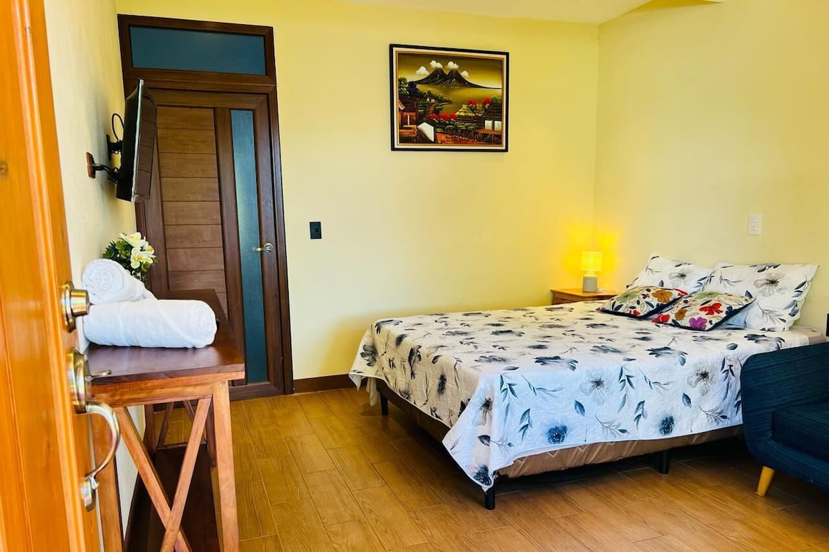 Un dormitorio con cama, mesita de noche con toalla, lámpara y un cuadro en la pared.