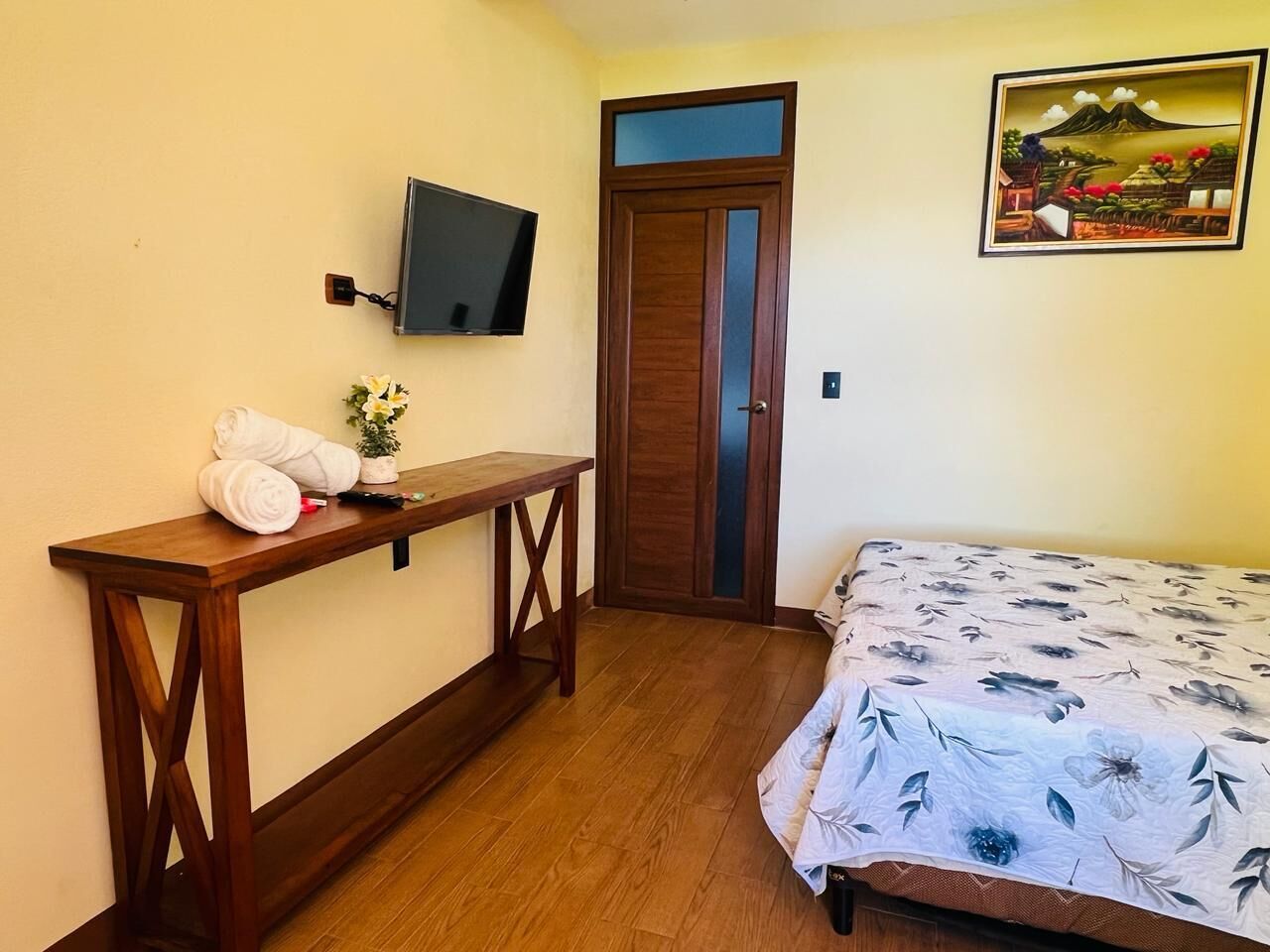 Una habitación con un escritorio de madera, una cama con colcha estampada, un televisor montado en la pared y un cuadro enmarco con un paisaje.