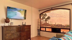 A living room with a TV, a wooden cabinet, a colorful sofa, and a large window with a view of palm trees and buildings. - Puerto Nuevo Villa Las Conchas 2BR 2 BA with pool (Centro Turístico El Pescador)