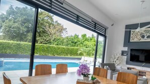 A modern dining area with a wooden table and chairs, a vase with flowers, and a view of a swimming pool and greenery outside. - Modern Mediterranean style house in Melgar condominium (Carmen de Apicala)