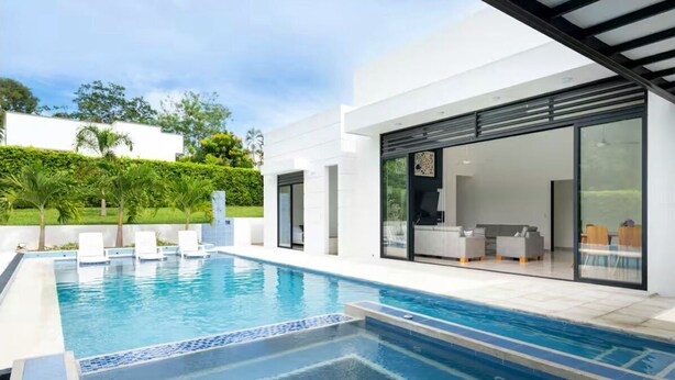 A modern outdoor swimming pool with a clear blue water feature, surrounded by a white building with large glass doors and a covered patio area. - Modern Mediterranean style house in Melgar condominium (Carmen de Apicala)
