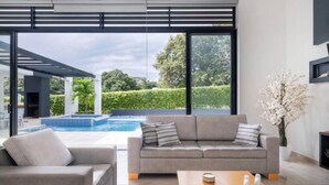 A modern living room with a grey sofa, a wooden coffee table, and a large window overlooking a swimming pool and greenery. - Modern Mediterranean style house in Melgar condominium (Carmen de Apicala)