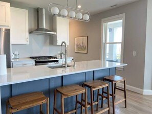 A modern kitchen with a large island, bar stools, stainless steel appliances, and a window providing natural light. - Premier Oceanfront Beach Pad 9BR (Atlantic City)