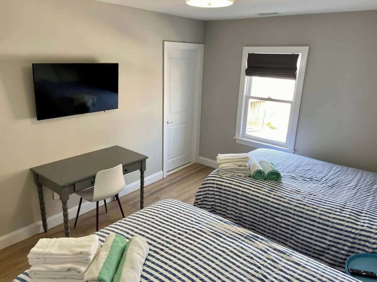 A room with two beds, a desk, a chair, a TV, and a window with blinds.