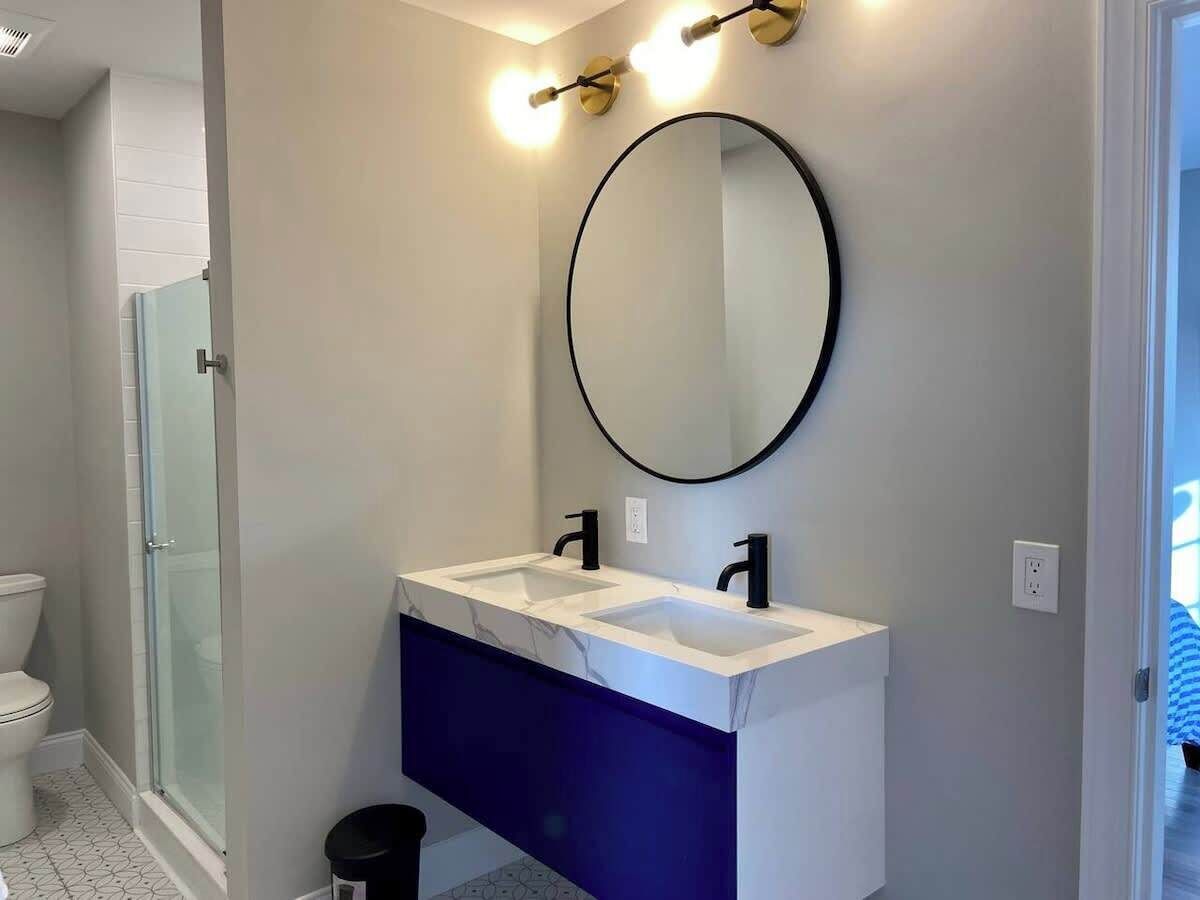 Un baño con cabina de ducha de vidrio, un lavabo blanco sobre un mueble de tocador azul y un espejo redondo con luces empotradas.