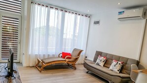 Una sala de estar con piso de madera, un sofá, una silla, un televisor sobre una mesa de madera y una ventana con cortinas.