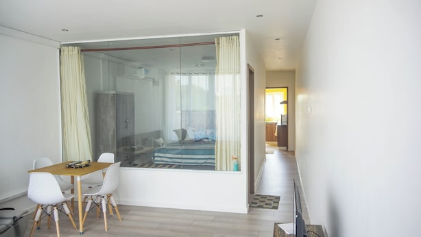 Una habitación moderna con un separador de vidrio, una cama, un área de comedor con mesa y sillas, y vistas a una cocina al fondo.