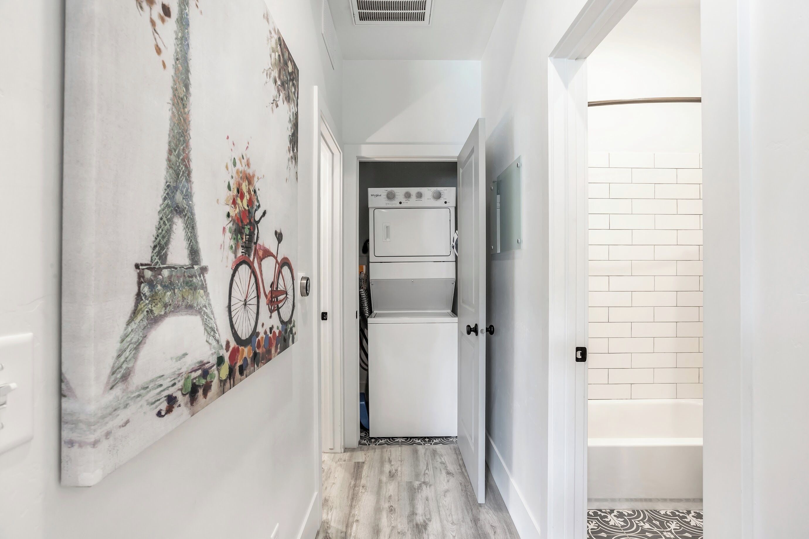 Passadís estret amb un frigorífic i un forn blancs, una porta que dona a un bany amb un mur de rajoles blanques, i una obra d'art penjada a la paret que representa una bicicleta i la Torre Eiffel.