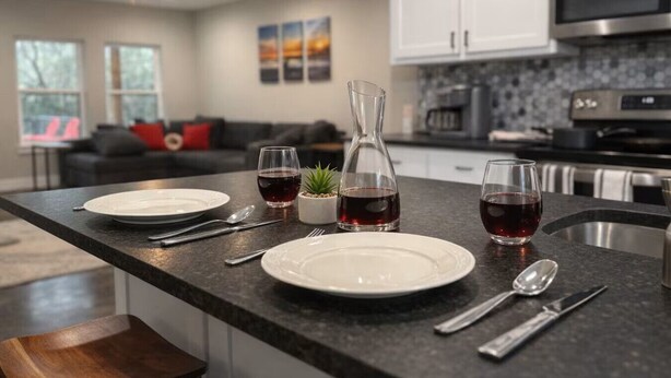 Una cocina moderna con encimera de granito oscuro, armarios blancos y electrodomésticos de acero inoxidable. Hay dos copas de vino y un decantador sobre la encimera.