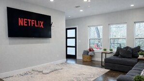 Una sala de estar moderna con un televisor de pantalla plana grande montado en la pared, un sofá gris, una mesa de centro y un ventanal con vistas a árboles.