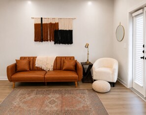 A living room with a brown sofa, a white armchair, a patterned rug, a small side table with a lamp, and a wall-mounted tapestry.