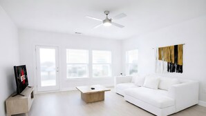 Un salon spacieux avec un canapé blanc, une table basse et un téléviseur posé sur un meuble. De grandes fenêtres sont équipées de stores, et le salon est agrémenté d’un ventilateur de plafond et d’une œuvre d’art accrochée au mur.