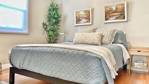 A bedroom with a bed, a bedside table, a window, and two framed pictures on the wall. - VIP YXE - Broadway |Three Bedroom Main Floor Suite (Saskatoon)