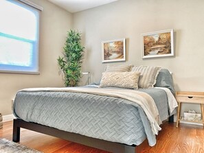 A bedroom with a bed, a bedside table, a window, and two framed pictures on the wall. - VIP YXE - Broadway |Three Bedroom Main Floor Suite (Saskatoon)