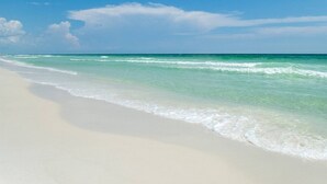 Une plage à l’eau turquoise et limpide, bordée de sable blanc, sous un ciel bleu.
