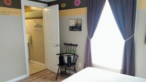 Ein Schlafzimmer mit einem Bett, einem Sessel mit Pflanzen, einer Tür zu einem Badezimmer und einem Fenster mit Vorhängen.