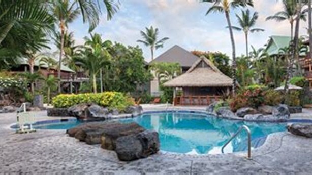 Un área de piscina en un resort con sillones, una estructura de techo de paja y vegetación tropical.