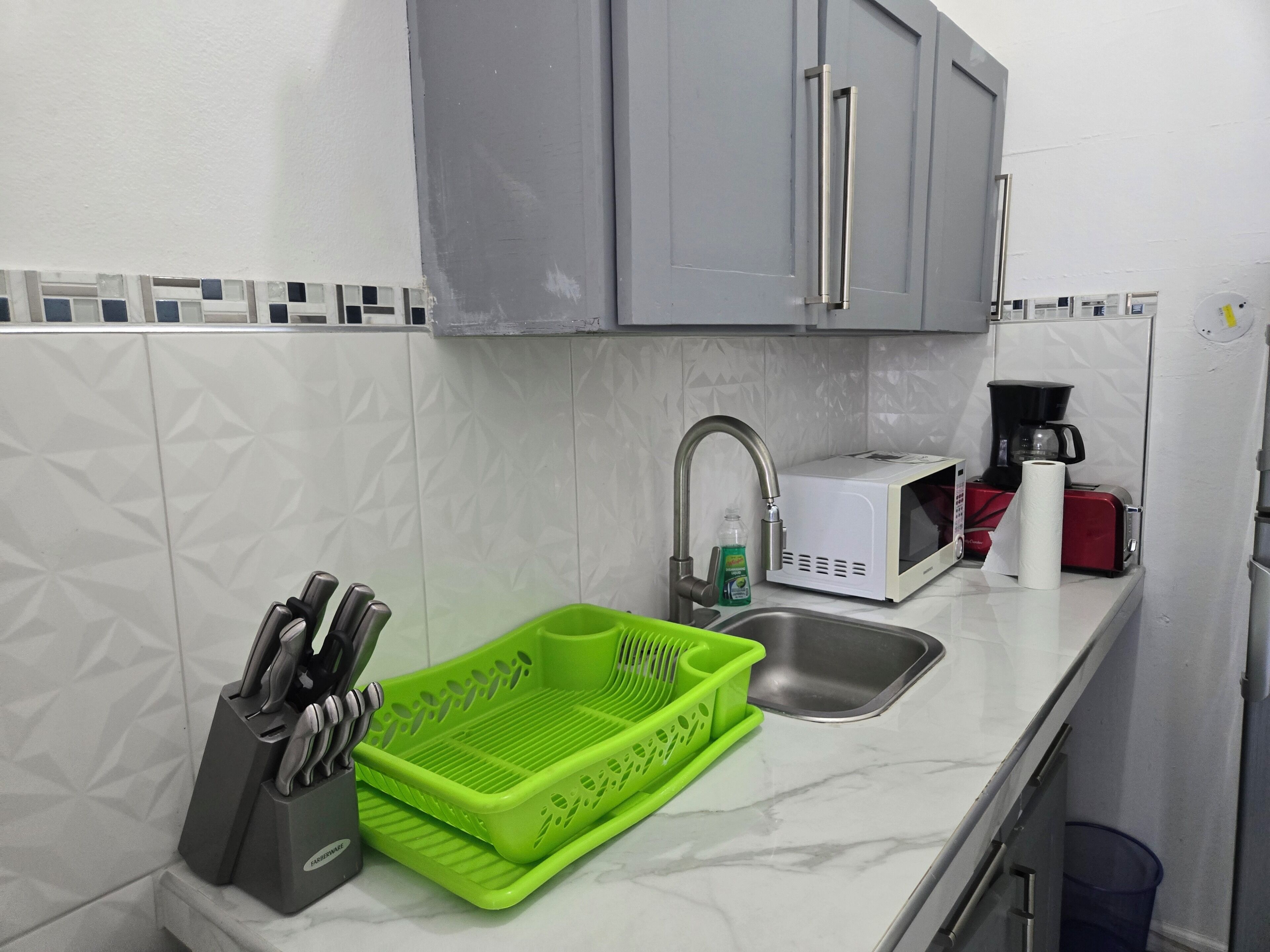 Une cuisine avec des meubles gris, un évier, un égouttoir à vaisselle vert, une machine à café et un grille-pain rouge.