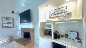 Un salon doté d’une cheminée, d’un téléviseur à écran plat fixé au-dessus, et d’un coin bar à café avec une enseigne indiquant « BAR À CAFÉ ».