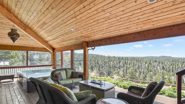 A covered outdoor area with a hot tub, wicker furniture, and a view of a forested landscape.