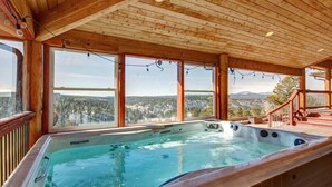 A wooden cabin with a hot tub and large windows overlooking a forested landscape.