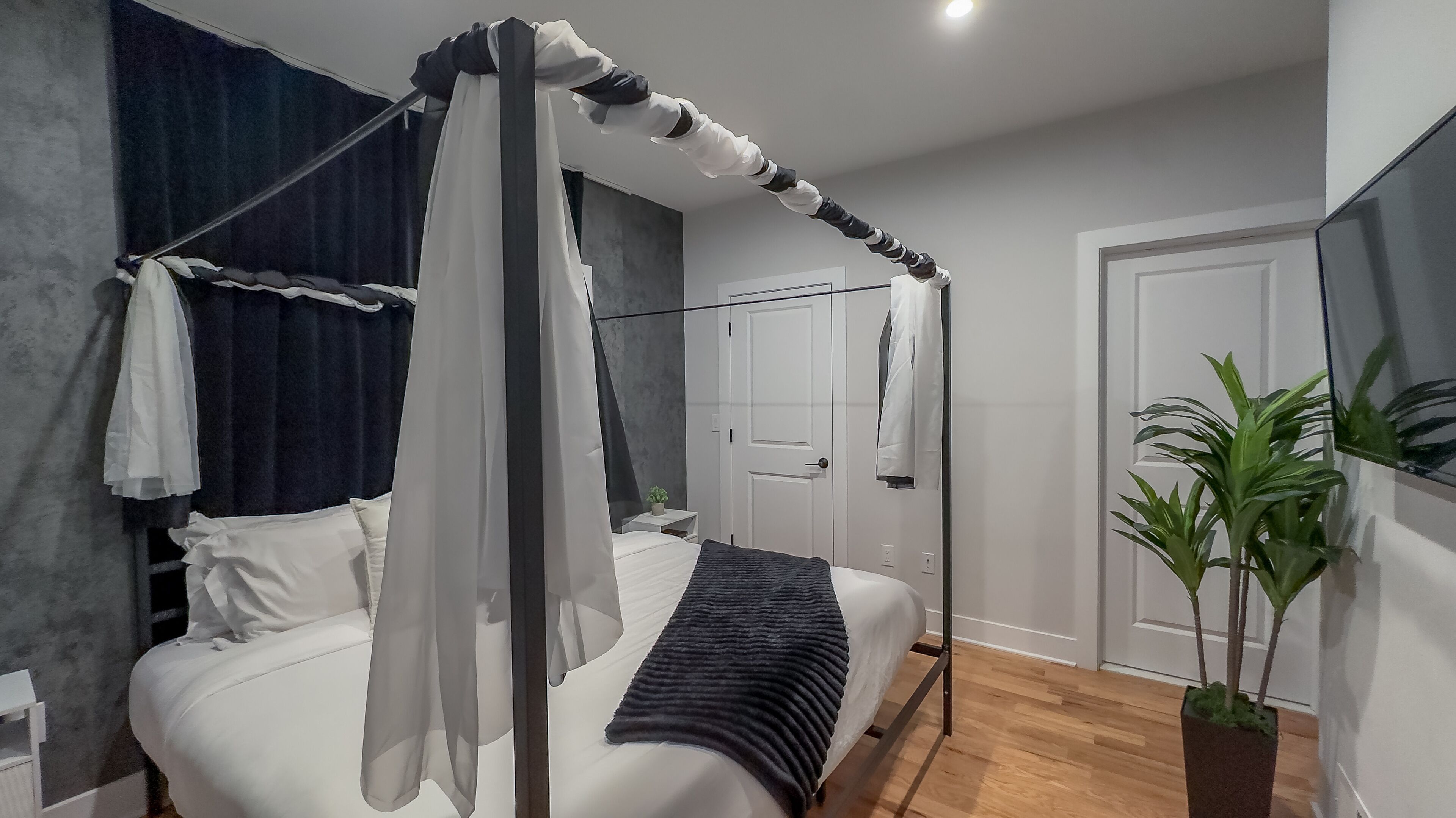 A modern bedroom with a four-poster bed, a bedside table, a nightstand, a potted plant, and a wardrobe.