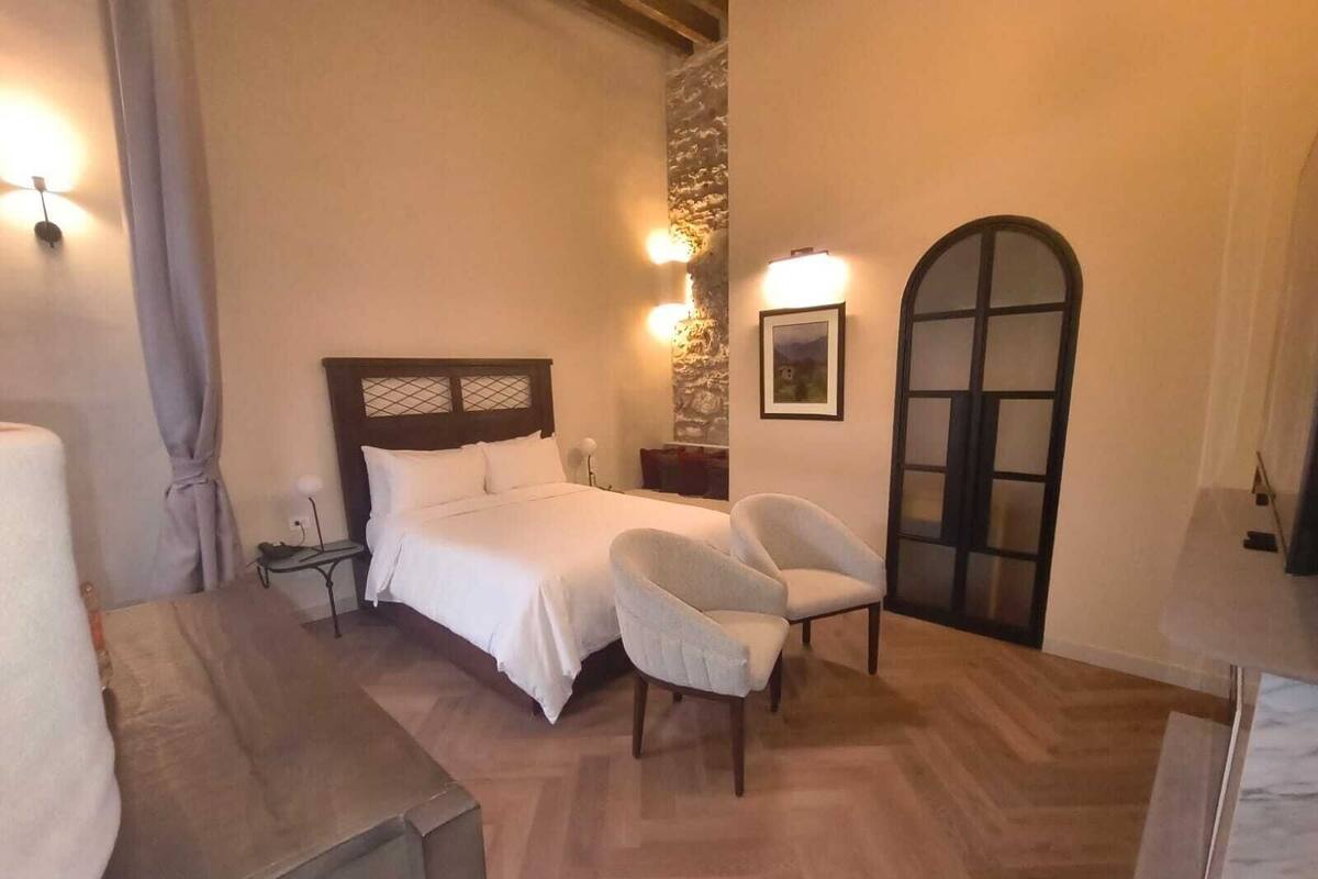 Ein Schlafzimmer mit einem Bett, zwei Stühlen, einem Nachttisch, einer Lampe, einem Bild an der Wand und einer Tür mit einem Fenster.