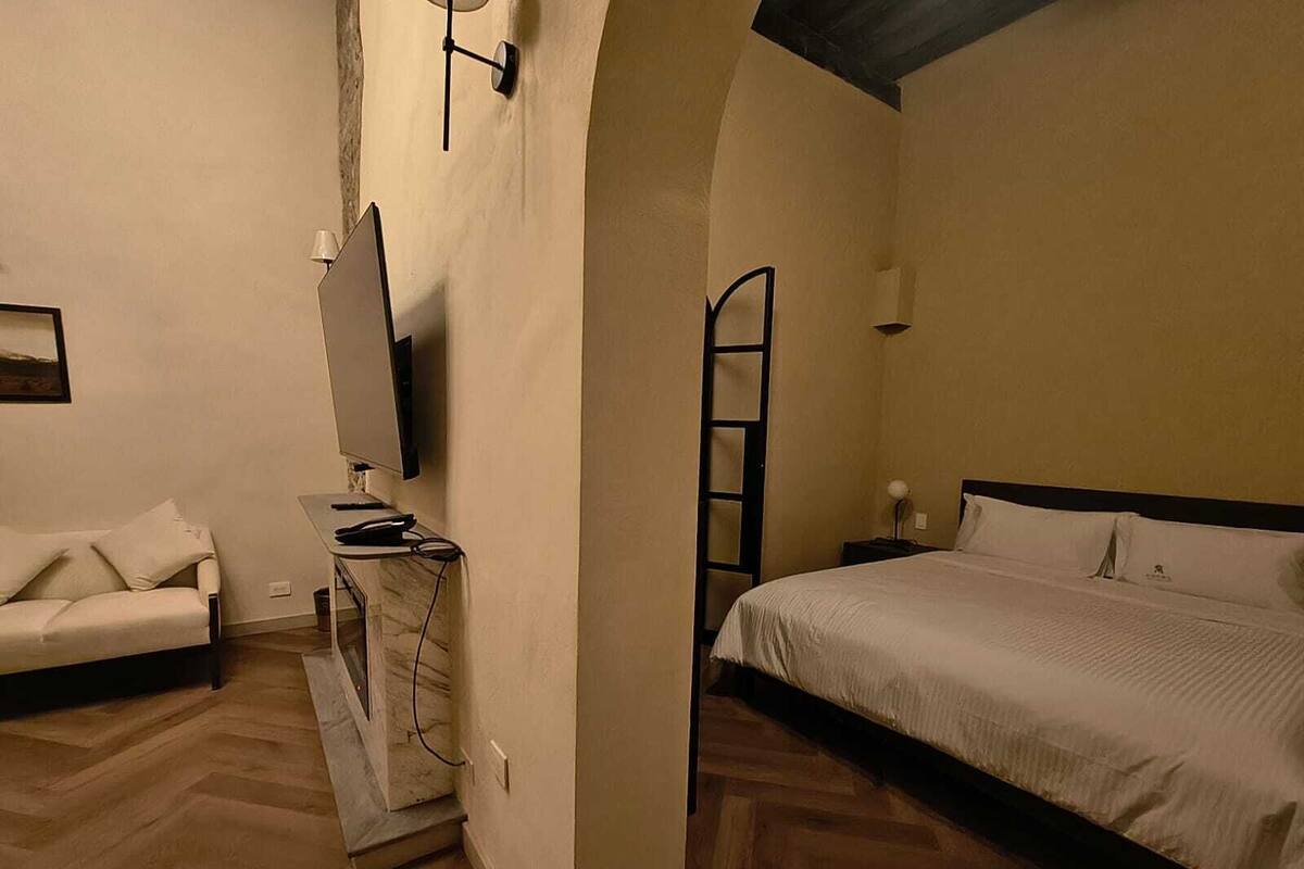 Ein Schlafzimmer mit einem Bett, einem Schreibtisch mit Computer, einem Sessel und einem an der Wand befestigten Fernseher.