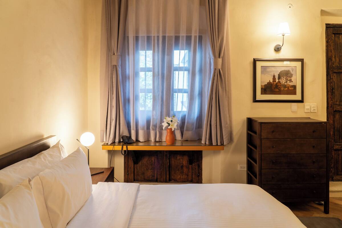 Ein Schlafzimmer mit Bett, Nachttisch mit Lampe, Kommode, Fenster mit Vorhängen und einem gerahmten Bild an der Wand.