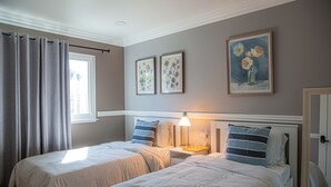 Ein Schlafzimmer mit zwei Betten, einem Fenster mit Vorhängen, einem Nachttisch mit Lampe sowie zwei gerahmten floralen Bildern an der Wand.