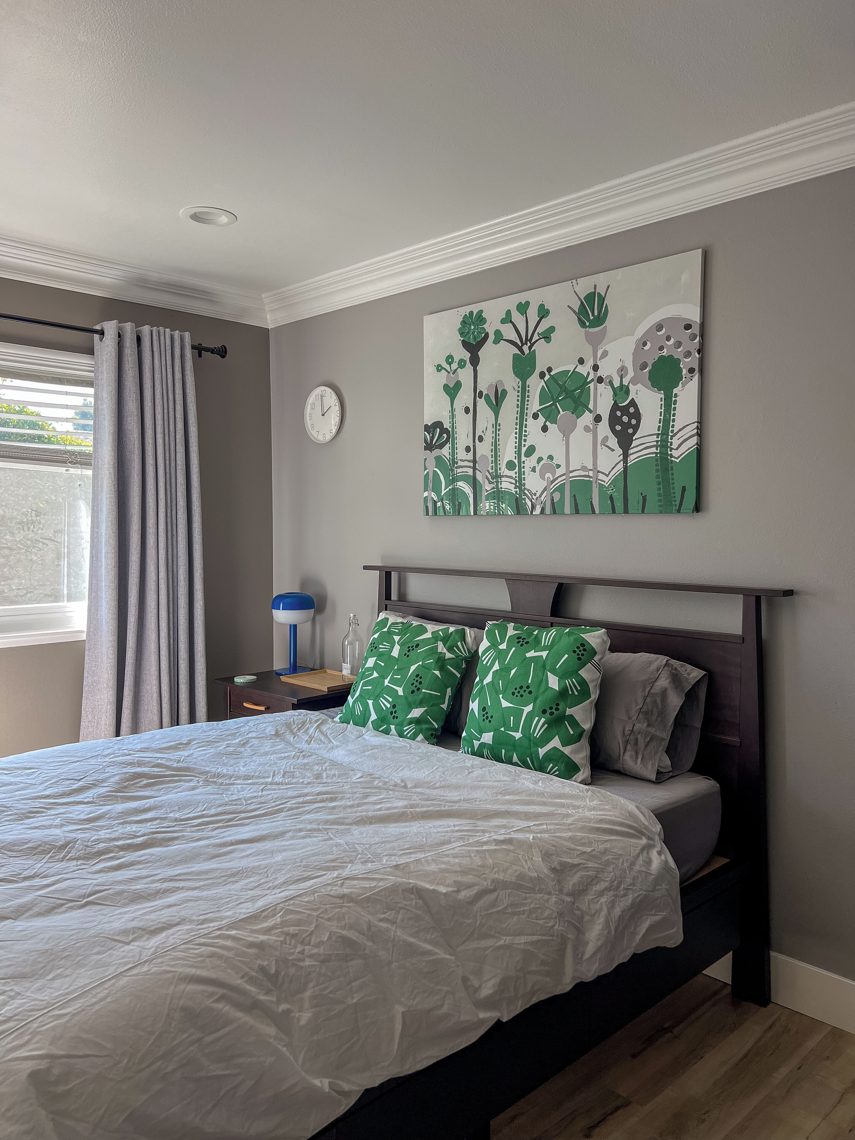 A bedroom with a bed, a clock, a painting, and a window with curtains.