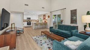 A modern living room with a teal sofa, a wooden coffee table, and a TV on a stand. There is a kitchen in the background with white cabinets and a dining area with a table and chairs. - Stylish Two Bedroom House in Peaceful Neighborhood (Carlsbad)