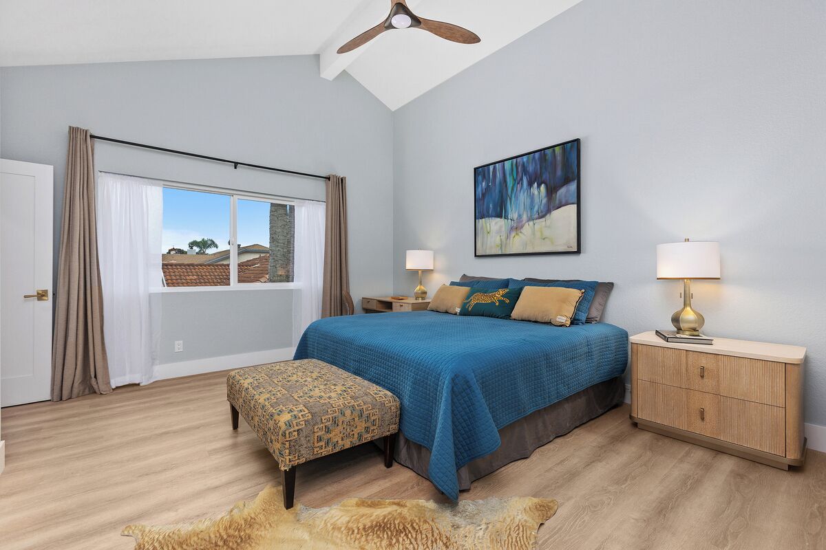 Une chambre avec un grand lit, un banc, une table de chevet, un ventilateur de plafond, une fenêtre avec des rideaux et un tableau au mur.