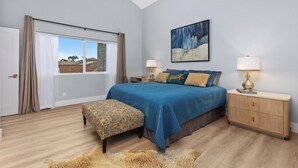 A bedroom with a large bed, a bench, a nightstand, a ceiling fan, a window with curtains, and a painting on the wall. - Stylish Two Bedroom House in Peaceful Neighborhood (Carlsbad)