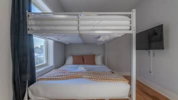 En våningssäng med vita sängkläder, en beige pläd och utsikt över staden genom ett fönster.