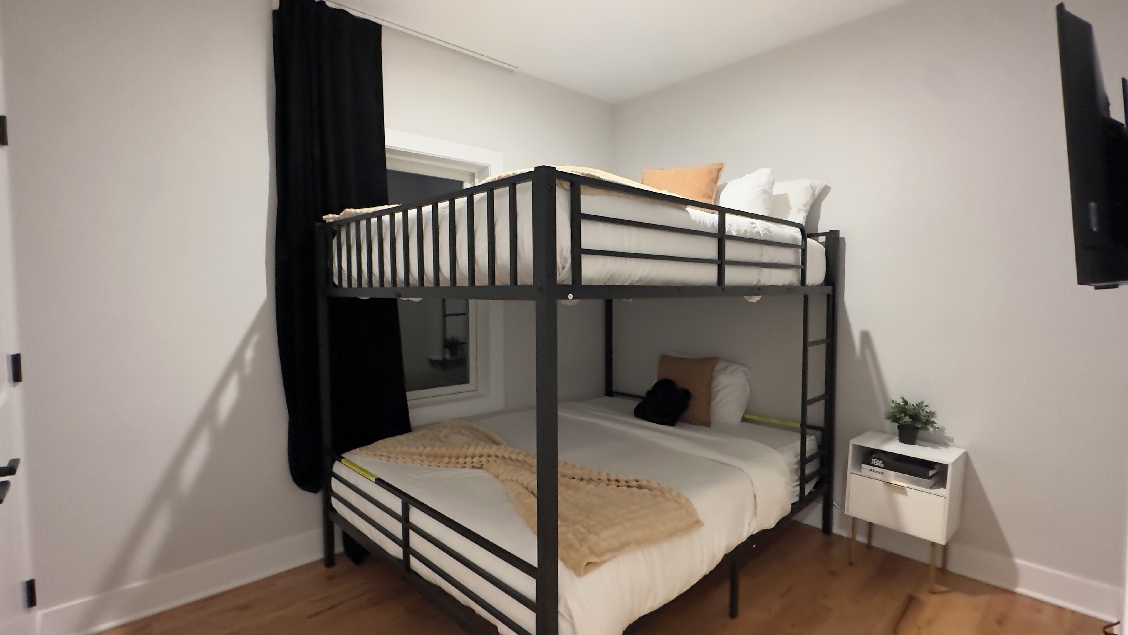 A bunk bed room with a window, a bedside table with a plant, and a wall-mounted TV.