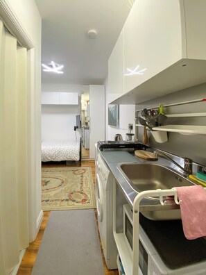 A compact kitchen with a stove, sink, and microwave. A bed and a small table are visible in the adjacent room. - Cosy Designed Studio in Willesden Green (London)