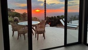 Een kamer met een glazen deur die uitkomt op een balkon met uitzicht op een zonsondergang boven een stadsgezicht.