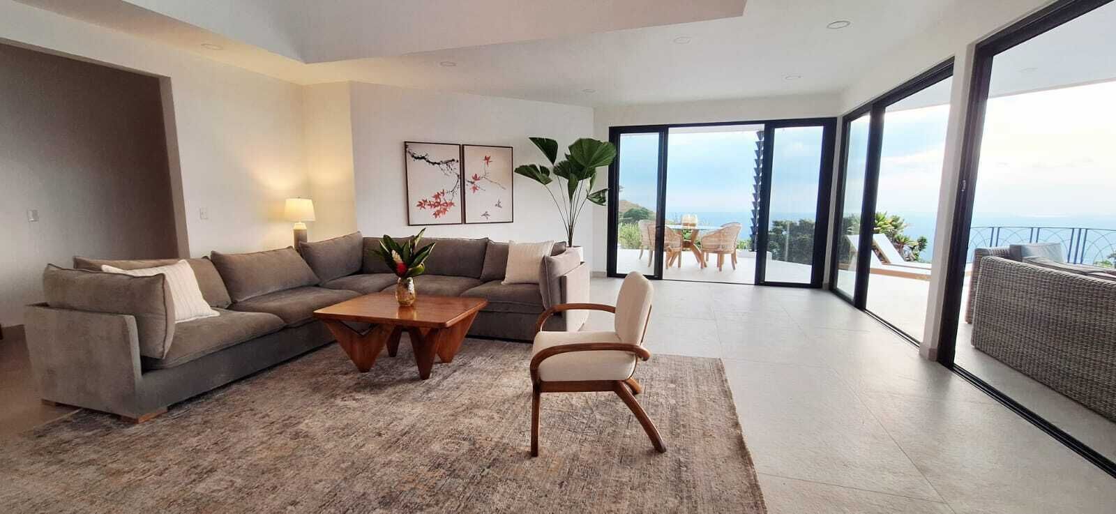 Un salon moderne avec un canapé, une table basse et un fauteuil, une grande fenêtre offrant une belle vue et une plante.
