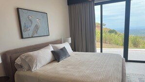 Een bed met een gestructureerd wit dekbed, twee kussens, een lijstwerk met een afbeelding van vogels, een lamp en een uitzicht op groen en een landschap in de verte door grote ramen.