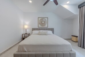 Un dormitorio con una cama, una mesita de noche, una lámpara, una obra de arte enmarcada y un ventilador de techo.