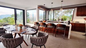 Een moderne eetruimte met een grote ronde tafel en meerdere stoelen, een keuken met houten keukenkastjes en grote ramen met uitzicht op de bergen.