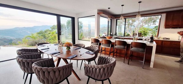 Un espace repas moderne avec une grande table ronde et de nombreuses chaises, une cuisine dotée de meubles en bois et de grandes fenêtres offrant une vue sur les montagnes.